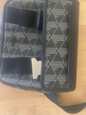 Svart axelväska från Lacoste - Snygg svart axelremsväska från Lacoste med geometriskt mönster och vita detaljer. Väskan har justerbar rem, metallspänne och dragkedja. Tillverkad i slitstarkt konstläder med tydlig Lacoste-logga på framsidan. har även påsen till väskan kvar!