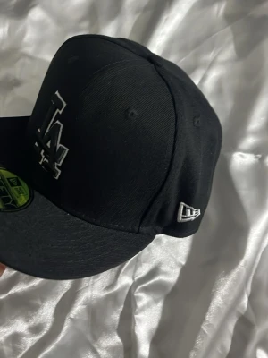Svart LA-keps från New Era - Snygg svart keps från New Era med LA-logga broderad i vitt framtill. Klassisk 59FIFTY-modell med rak skärm och broderad New Era-logga på sidan. Tillverkad i bomullsmaterial och passar perfekt för en streetwear-look.