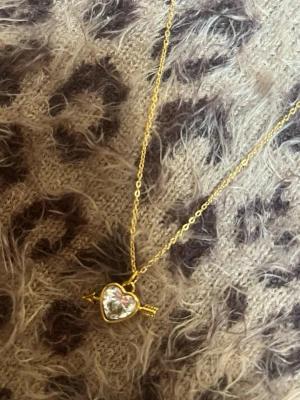 Halsband med hjärthänge och pil guld vit  - Guldfärgat halsband med ett hjärta i klar sten och en liten pil genom hänget. Kedjan är tunn och elegant, och hänget har en glittrande, kristalliknande sten i mitten. Perfekt accessoar för en trendig look. Rostfri 