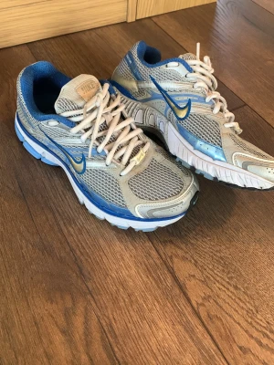 Nike blå och grå sneakers structure retro klassiska  - Snygga sneakers från Nike i grått och blått med vita detaljer. Skorna har mesh-ovandel för bra ventilation, vita snören och en blå swoosh på sidan. Sulan är vit med blå inslag och insidan är vadderad för extra komfort. Perfekta för dig som gillar sportig stil.