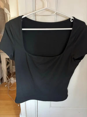 Svart topp med fyrkantig ringning - En stilren svart topp med korta ärmar och fyrkantig ringning. Toppen har en tight passform och är tillverkad i ett mjukt, stretchigt material som sitter snyggt på kroppen. Perfekt att matcha med jeans eller kjol för en trendig look.