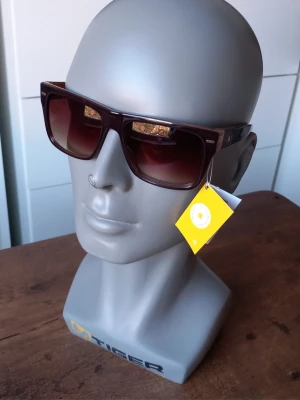 Bruna solglasögon från Sunshine  - Snygga solglasögon från Sunshine med bred båge i mörkbrunt och röda detaljer på skalmarna. Klassisk rektangulär form som ger en cool vibe och passar till de flesta outfits. Perfekta för soliga dagar när du vill sticka ut lite extra.