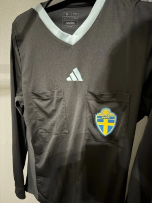 Svart domartröja Adidas SvFF - Svart långärmad domartröja från Adidas med två bröstfickor och SvFF-emblem på vänster sida. Tröjan har ljusgrå V-ringning och Adidas-logga på bröstet. Perfekt för fotbollsdomare. Tillverkad i polyester för sportig känsla.