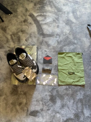 Valentino rockrunners grå/svart - Snygga sneakers från Valentino i grått, svart och vitt camomönster med röda detaljer på hälen. Skorna har lågt skaft, mesh och mocka-paneler samt svarta sulor. Kommer med originalkartong, dustbag och extra skosnören. Perfekta för dig som gillar streetstyle och exklusiva märken.