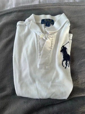 Vit piké från Polo Ralph Lauren - S i barnstorlek! Klassisk vit pikétröja från Polo Ralph Lauren med ikonisk mörkblå broderad logga på bröstet. Skjortan har krage, knappar framtill och är gjord i mjuk bomull. Perfekt för en clean och stilren look.