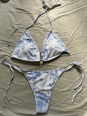 Bikini - Snygg bikini i vitt med blått blommönster. Triangelformad topp med knyt i nacken och ryggen samt matchande trosa med knyt i sidorna. Perfekt för stranden eller poolen, ger en fräsch och somrig look. Helt oanvänd! Säljer pga att jag råkade beställa två likadana!