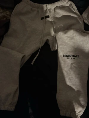 Gråa Essentials mjukisbyxor - Snygga grå mjukisbyxor från Fear of God Essentials med logga tryckt på benet och svart patch framtill. Byxorna har dragsko i midjan och resår vid benslut. Perfekta för en chill och trendig look.