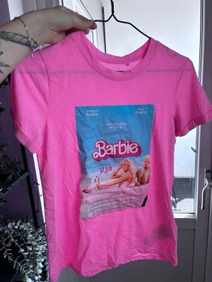 Barbie rosa t-shirt med tryck - Cool rosa t-shirt med Barbie-tryck framtill, inspirerad av filmen. T-shirten har rund hals och korta ärmar. Perfekt för dig som älskar färg och popkultur. Materialet känns mjukt och skönt mot huden. T-Shirten är stor i storleken och passar även om man är storlek Small. 