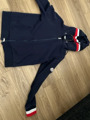 Marinblå Moncler hoodie med dragkedja - Snygg marinblå hoodie från Moncler med dragkedja framtill och huva. Detaljer i rött och vitt på huvan och ärmslut samt Moncler-logga på ärmen. Tillverkad i mjukt material och har fickor framtill. Perfekt för en sportig och stilren look.