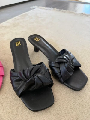Svarta slip-in sandaler med klack dinsko - Stilrena svarta sandaler från dinsko med låg klack och fyrkantig tå. Ovandelen har en snygg knutdetalj i vadderat konstläder som ger en trendig look. Perfekta för dig som vill ha en chic och bekväm sko till sommaren.