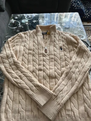 Beige kabelstickad tröja Polo Ralph Lauren - Kabelstickad beige tröja från Polo Ralph Lauren med halv dragkedja och ribbade muddar. Tröjan har en klassisk design med broderad logga på bröstet och är tillverkad i mjukt material. Perfekt för en stilren och avslappnad look.