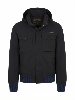 Svart vindjacka från Woolrich - Snygg svart vindjacka från Woolrich med huva, två stora bröstfickor med knappar och en dragkedjeficka. Jackan har ribbade muddar i mörkblått vid ärmslut och nederkant samt diskret logotyp på ärmen. Tillverkad i slitstarkt material, perfekt för blåsiga dagar.