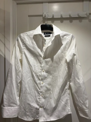 Vit skjorta från Riley | Strl S - Klassisk vit skjorta från Riley i slim fit-modell. Skjortan har långa ärmar, knappar framtill och en stilren krage. Perfekt för dig som gillar en clean och tidlös look. Tillverkad i bomull för en skön känsla.