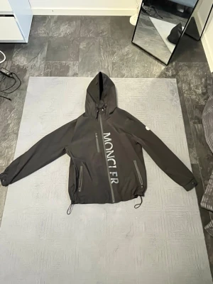 Svart Moncler vindjacka med huva - Svart vindjacka från Moncler med stor logga längs dragkedjan framtill. Jackan har huva, två snedställda fickor med dragkedja och justerbar dragsko i nederkant. Perfekt för blåsiga dagar och snygg streetstil.