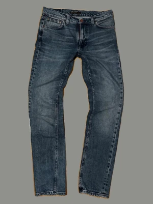 Blå jeans från Nudie - Snygga blå jeans från Nudie med klassisk femficksdesign och subtila slitningar. Jeansen har Nudies ikoniska broderi på bakfickorna och är tillverkade i mjuk denim med lite stretch för extra komfort.
