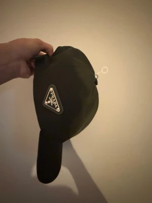 Svart keps från Prada i nylon - Snygg svart keps från Prada med det klassiska trekantiga metallogot på sidan. Kepsen är gjord i ett glansigt nylonmaterial och har en böjd skärm. Perfekt accessoar för en trendig och sportig look.