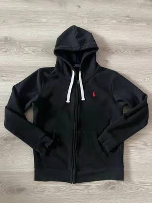 Svart hoodie från Polo Ralph Lauren - Svart hoodie med dragkedja från Polo Ralph Lauren. Klassisk design med vit dragsko i huvan och röd broderad logga på bröstet. Mjuk insida och känguruficka framtill. Perfekt för en chill och stilren look. Storlek M helt ny använd 1 gång