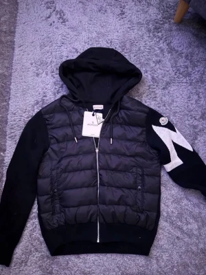 Moncler cardigan - Svart cardigan från Moncler med luva och dragkedja framtill. Jackan har quiltad front, stickade ärmar med vit zigzag-detalj och Moncler-logga på ena ärmen. Fickor med knappstängning och ribbade muddar. Materialmix av dun och bomull.