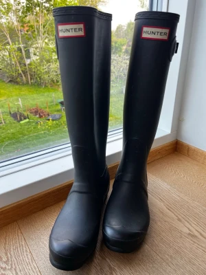 Svarta Hunter höga gummistövlar - Klassiska svarta höga gummistövlar från Hunter med justerbart spänne på sidan och logotyp framtill. Stövlarna är tillverkade i slitstarkt gummi och har en tidlös, stilren design som passar perfekt för regniga dagar. Varsamt använda.