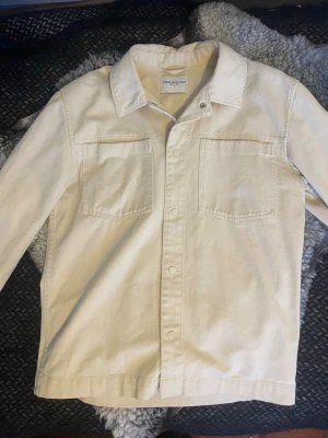 Beige overshirt från These Glory Days - Stilren beige overshirt från These Glory Days med klassisk krage och två stora bröstfickor. Overshirten har raka linjer, knäppning framtill och är tillverkad i ett kraftigare bomullstyg. Perfekt för lager-på-lager och enkel att matcha.