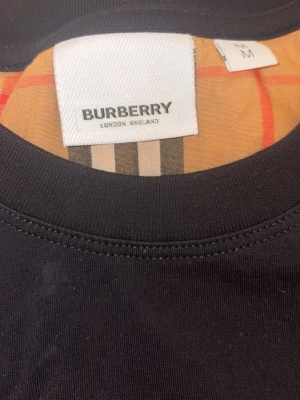 Burberry svart t-shirt med rutiga ärmar - Svart t-shirt från Burberry med klassiska beige-rutiga ärmar i signaturmönster. T-shirten har rund halsringning och en stilren passform som ger en snygg kontrast mellan svart och det ikoniska Burberry-mönstret.