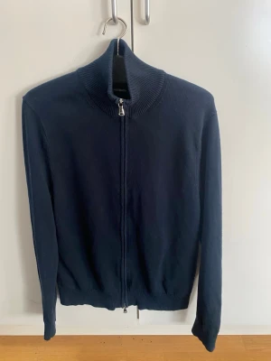 Fullzip cardigan J.Lindeberg - Mörkblå stickad ziptröja från J.Lindeberg med hög krage och hel dragkedja framtill. Tröjan har långa ärmar och ribbade muddar vid ärmslut och nederkant. Enkel och stilren design som passar till många olika outfits. Ny pris 1700kr. Storlek M.