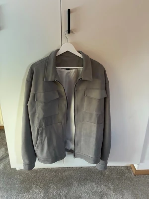 Grå overshirt med fickor - Snygg grå overshirt med klassisk krage och fyra stora fickor framtill. Jackan har dragkedja och är tillverkad i ett mjukt material som ger en avslappnad look. Perfekt att slänga över en t-shirt för en stilren outfit. Storlek M, passar dig som är 180-185cm lång och väger mellan 65-75 kg