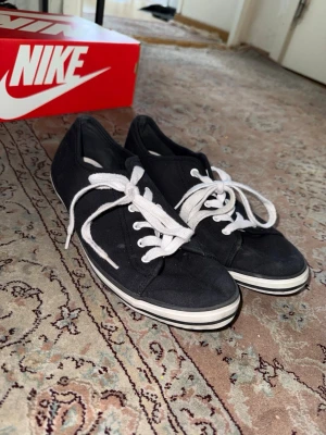 Svarta Nike canvas sneakers med vita snören 41/42 storlek  - Svarta låga sneakers från Nike i klassisk canvas med vita snören och vit sula med svart rand. Perfekta för en avslappnad och stilren look. Tidlös design som funkar till det mesta och är lätta att matcha.
