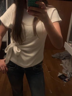Vit topp med knytdetalj i sidan - Snygg vit topp med korta ärmar och en trendig knytdetalj i sidan. T-shirten har en avslappnad passform och är gjord i ett mjukt, stretchigt material. Super bra skick! Använd cirka 1-2 gånger endast ❤️ 