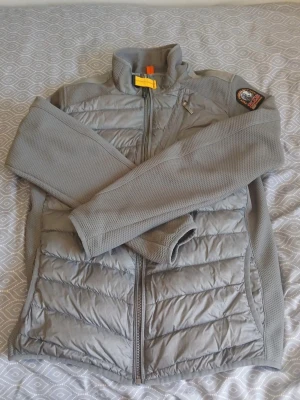 Grå hybridjacka från Parajumpers - Snygg grå jacka från Parajumpers med quiltad front och ärmar i stickad polyester. Jackan har dragkedja, hög krage och en bröstficka med dragkedja. Märkets patch på ärmen ger en sportig vibe.