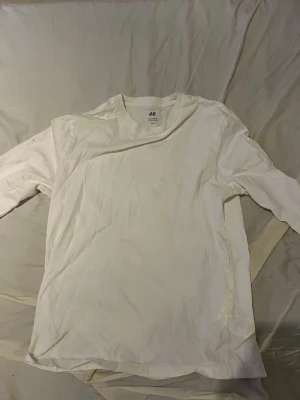 Vit långärmad t-shirt från H&M - En enkel och stilren vit långärmad t-shirt från H&M. Tillverkad i mjukt bomullsmaterial med rund halsringning och rak passform. Perfekt basplagg för alla tillfällen och lätt att matcha med andra plagg.