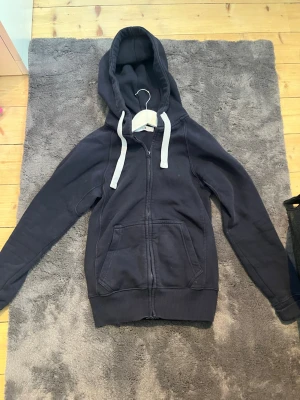 Mörkblå zip hoodie med vita snören - Mörkblå hoodie med dragkedja och stora fickor framtill. Har vita, breda dragsnören i huvan och ribbade muddar vid ärmar och nederkant. Perfekt för en avslappnad och chill stil. Materialet känns mjukt och bekvämt, troligen bomullsblandning.