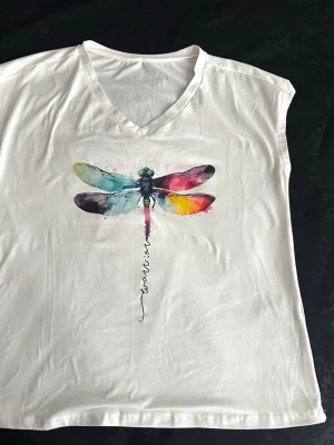 Vit t-shirt med färgglad trollslända M , B 52/L 62 - Vit t-shirt med v-ringning och ett stort tryck av en färgglad trollslända på bröstet. T-shirten är ärmlös och har en avslappnad passform. Perfekt för dig som gillar unika och kreativa motiv.