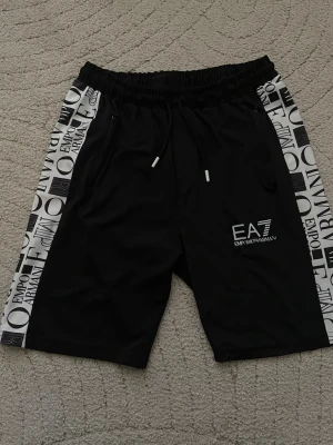 Svarta EA7 shorts med logga - Svarta shorts från Emporio Armani EA7 med vita sidopaneler med tryckt logomönster. Shortsen har snörning i midjan och en vit EA7-logga framtill. Perfekta för en sportig och trendig look.