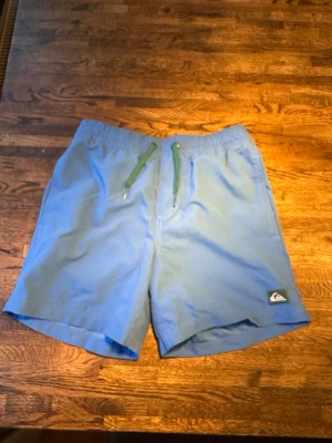 Blå badbyxor från Quiksilver juniormodell  - Säljer ett par ljusblå badbyxor från Quiksilver med elastisk midja och gröna snören. De har en bakficka med knapp och klassisk Quiksilver-logga framtill. Perfekta för stranden eller poolen i sommar!