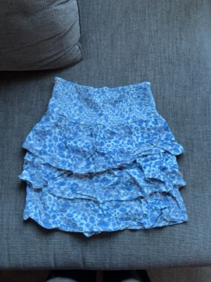 Blå blommig volangkjol - Söt kort kjol med blått blommönster och flera volanger. Kjolen har en bred resår i midjan och är gjord i ett lätt och luftigt material, perfekt för varma dagar. Passar dig som gillar en feminin och trendig stil. Typ aldrig använd