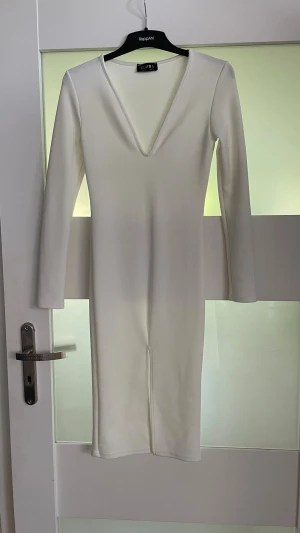 Vit långärmad klänning från Club L - Stilren vit klänning från Club L med lång ärm och djup v-ringning. Klänningen är figurnära och har en enkel, elegant design som passar perfekt till olika tillfällen. Tillverkad i ett stretchigt material för bekväm passform.