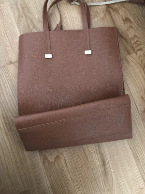 Brun toteväska i läderimitation - Stilren brun toteväska i läderimitation med två handtag och guldfärgade detaljer. Väskan har en rymlig insida med en innerficka med dragkedja och matchande clutch. Perfekt för dig som gillar klassisk och enkel design.