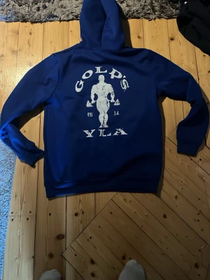 Blå Gold's Gym hoodie med tryck - Snygg blå hoodie från Gold's Gym med stort vitt tryck av en bodybuilder och text på ryggen samt mindre logga på bröstet. Klassisk huva med dragsko och magficka framtill. Perfekt för gymmet eller chill. Mjuk och bekväm bomullsblandning.