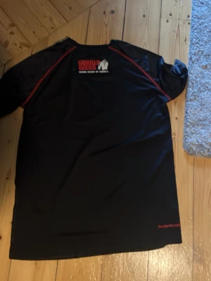 Svart funktions-tshirt Gorilla Wear - Säljer en svart funktions-tshirt från Gorilla Wear med röda sömmar och vit gorilla-logga på bröstet. Tillverkad i ett lätt och snabbtorkande material som passar perfekt till träning och gym. Klassisk rund hals och korta ärmar för maximal rörelsefrihet.