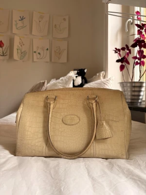 Beige handväska i krokodilmönster - Säljer en beige handväska från Mulberry med krokodilmönstrad struktur och klassisk oval logga framtill. Väskan har två rejäla handtag och ett dekorativt hänglås. Insidan är fodrad med rutigt tyg och har flera fack för smart förvaring.