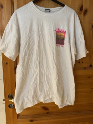 Vit Stüssy t-shirt med tryck - Vit t-shirt från Stüssy med färgglatt tryck på både bröstet och ryggen. T-shirten har en klassisk passform och rund halsringning. Materialet är mjuk bomull och trycket har inslag av rosa, gult och svart.