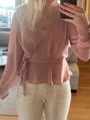 Rosa omlottblus med knytband - Söt rosa omlottblus med knytband i midjan och volang nertill. Blusen har lång ärm och v-ringad halsringning. Perfekt för en trendig och feminin look. Materialet ser ut att vara mjukt och lätt, vilket ger en skön känsla.