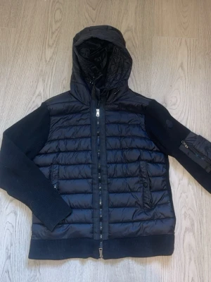 Svart pufferjacka från Moncler - Snygg svart pufferjacka från Moncler med stickade ärmar och huva. Jackan har quiltad front, två fickor med dragkedja och en extra ficka på ärmen. Moncler-logga på vänster ärm. Perfekt för kalla vinterdagar.
