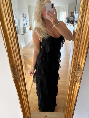 Svart långklänning med volanger - Säljer en svart långklänning med volanger i flera lager och smala axelband. Klänningen har en elegant och flowy siluett som ger en cool look. Perfekt för dig som vill sticka ut med en klassisk men trendig stil.