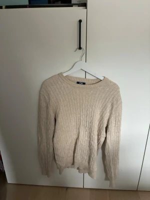 Beige kabelstickad tröja från Cutter & buck - Kabelstickad tröja i beige med rund halsringning och ribbade muddar. Tröjan har en klassisk passform och är perfekt för lager-på-lager. Perfekt till sena sommarkvällar! Nypris 1100!