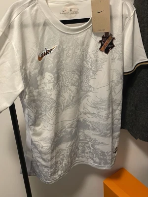 Vit AIK x Nike matchtröja - Säljer en vit AIK matchtröja från Nike med detaljer i guld och svart. Tröjan har ett unikt tryck med Stockholmsmotiv, AIK:s klubbmärke på bröstet och Nike-logga i guld. Tillverkad i lätt och ventilerande polyester, perfekt för träning eller match.