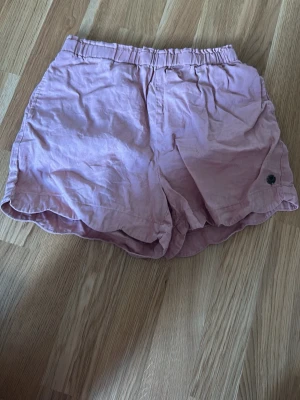 shorts med resårmidja - Säljer ett par aprikos färgade shorts med elastisk midja och avslappnad passform. Shortsen har en mjuk känsla och är perfekta för varma dagar. De har en liten logga på ena benet och är tillverkade i ett lätt material.