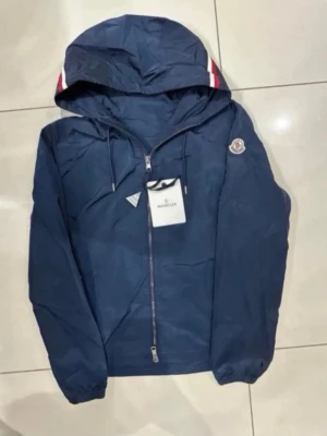 Moncler Vindjacka - Snygg marinblå vindjacka från Moncler med huva och dragkedja framtill. Jackan har Moncler-logga på ärmen och vita/röda ränder på huvan. Lätt material, perfekt för blåsiga dagar. Justerbar dragsko i huvan och elastiska muddar vid ärmslut.