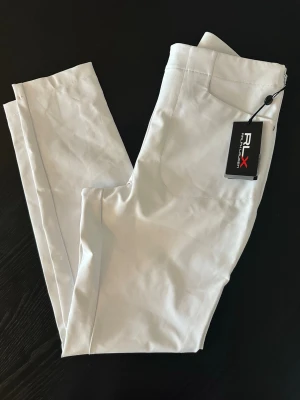  RLX Ralph Lauren vita byxor - Snygga vita byxor från RLX Ralph Lauren med raka ben och klassisk design. Byxorna har diskreta fickor och är tillverkade i ett mjukt och stretchigt material som ger en bekväm passform. Perfekta för en clean och stilren look.